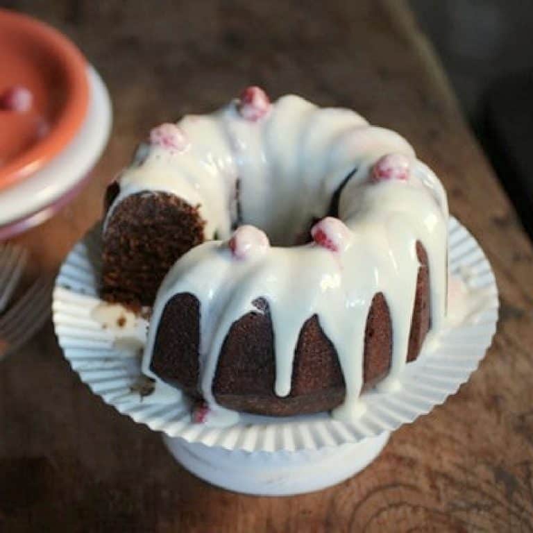 White glaze over chocolate bundt cake on a white cake stand on a wooden table.