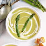 Close up white bowl of asparagus soup. White surface, silver spoons, fresh asparagus.