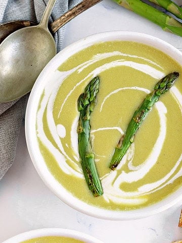 Close up white bowl of asparagus soup. White surface, silver spoons, fresh asparagus.