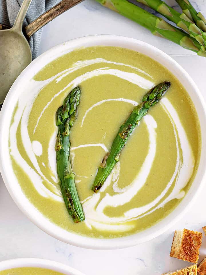 Close up white bowl of asparagus soup. White surface, silver spoons, fresh asparagus.