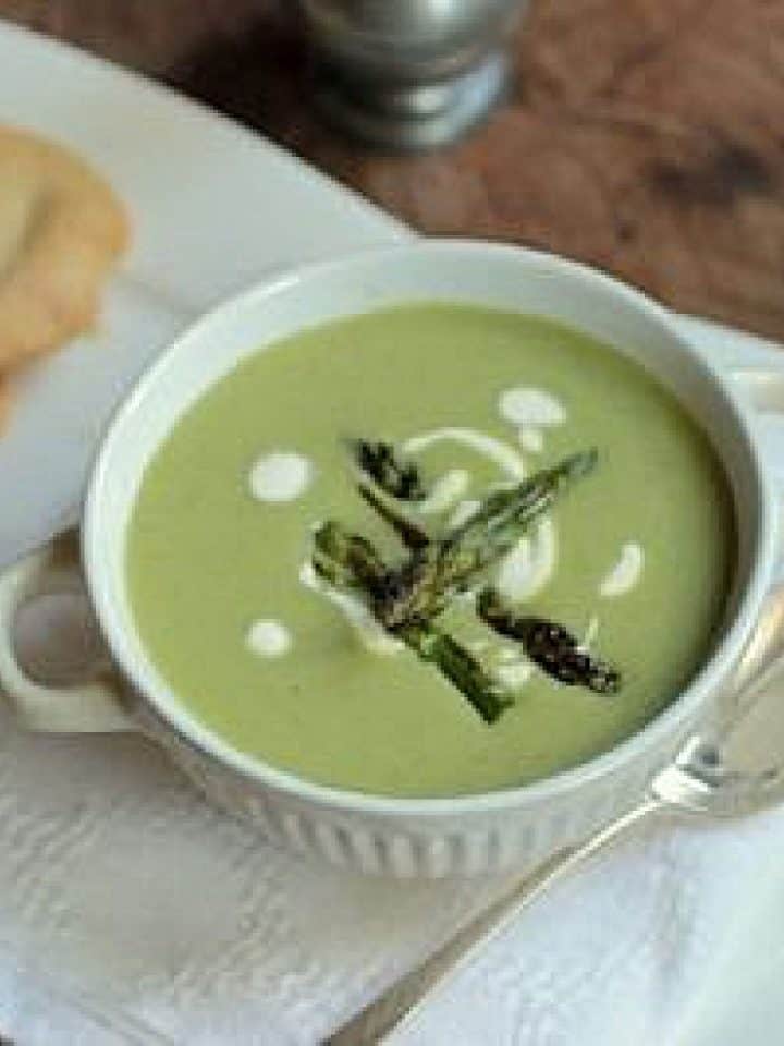 Top view of white bowl with green asparagus soup on a white napkin. A silver spoon and crackers around.