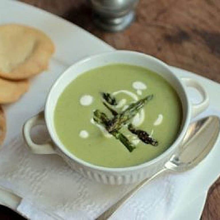 Top view of white bowl with green asparagus soup on a white napkin. A silver spoon and crackers around.