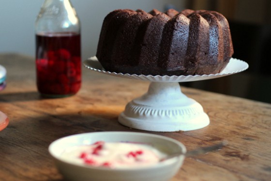 Chocolate bundt cake on white stand, bowl and bottle with raspberries, wooden table