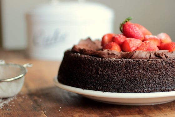 Partial view of chocolate cake with strawberries, wooden table, white prop in background