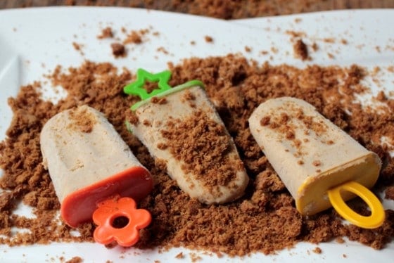 Three cinnamon ice cream popsicles with colorful holders on a white plate with graham cracker crumbs. 