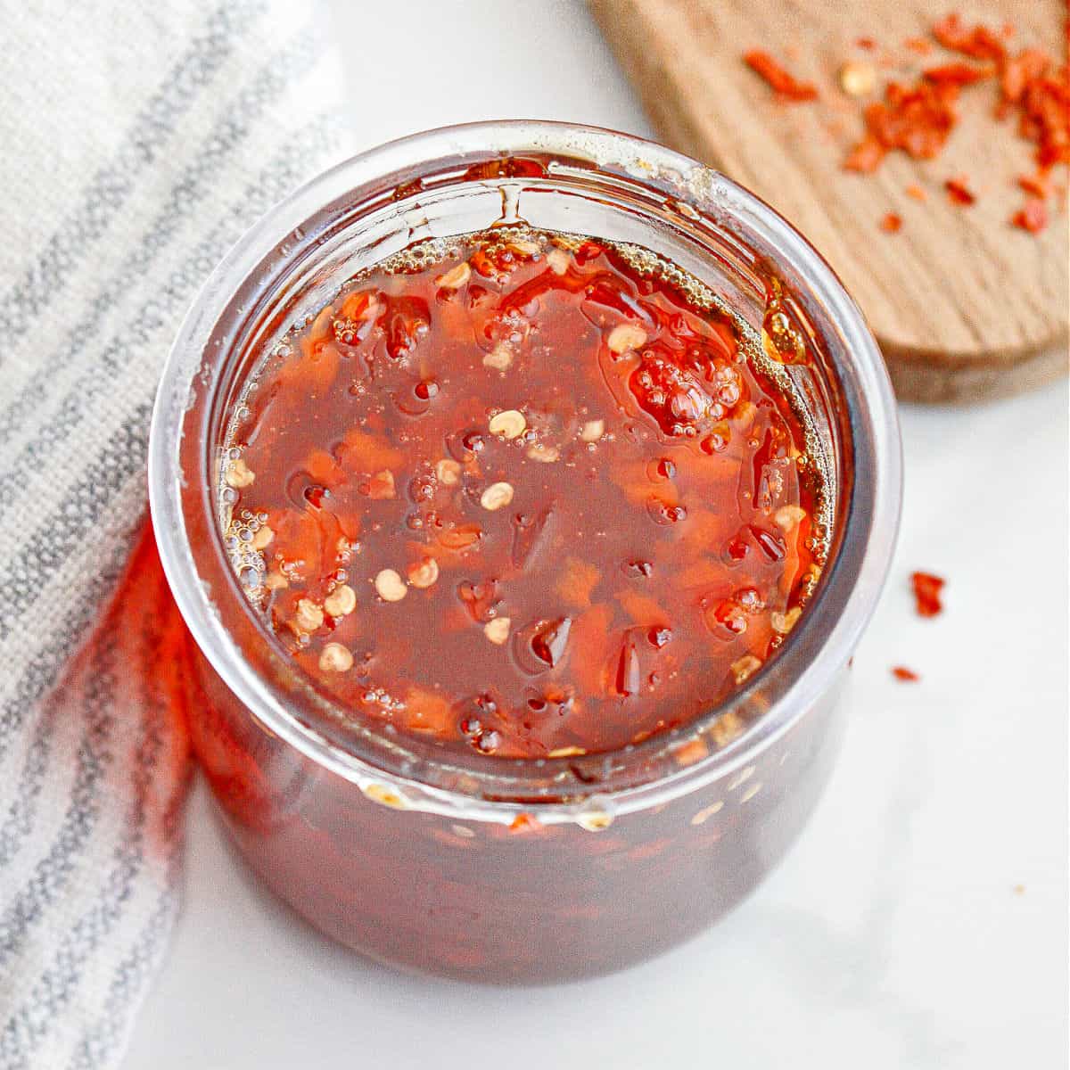Top view of jar with red chili jam. White surface, striped cloth. 