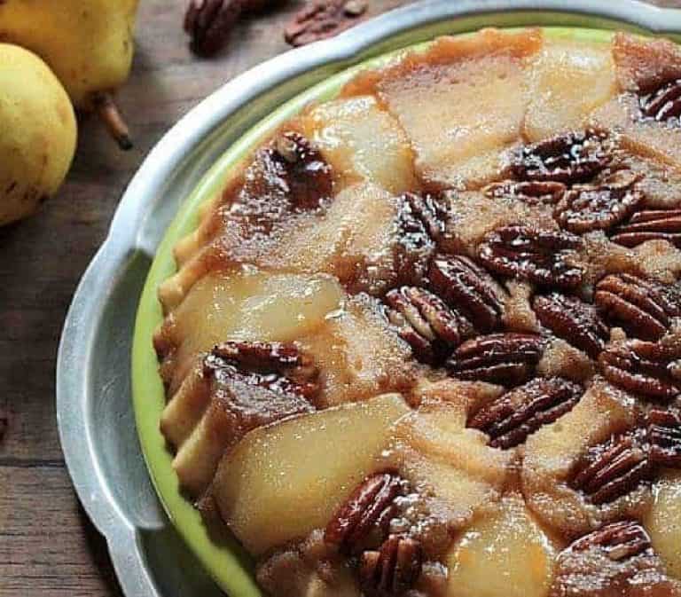 Partial pear upside down cake on green and silver platter; wooden table, whole pears, scattered pecans