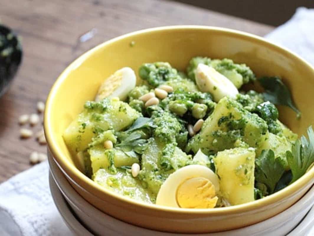 Close up of potato Pesto Egg Salad in yellow bowl on a white linen and wooden table.