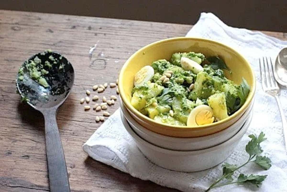 Top view of potato egg salad with pesto in a yellow bowl stack and white linen. Wooden table with wooden spoon. 