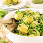 Potato pesto spinach salad in a white bowl with wooden tongs. Plates and bowl with pesto and potatoes in the background.
