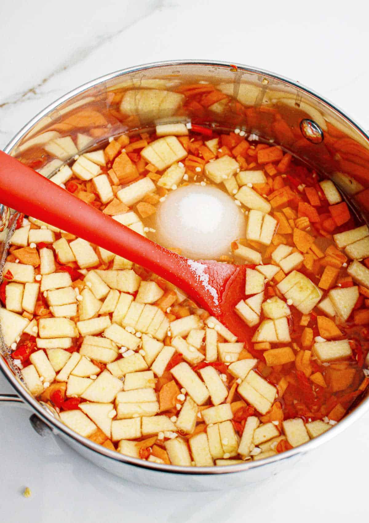 Large pot with ingredients for red chili jam with apples, sugar and vinegar. White surface.