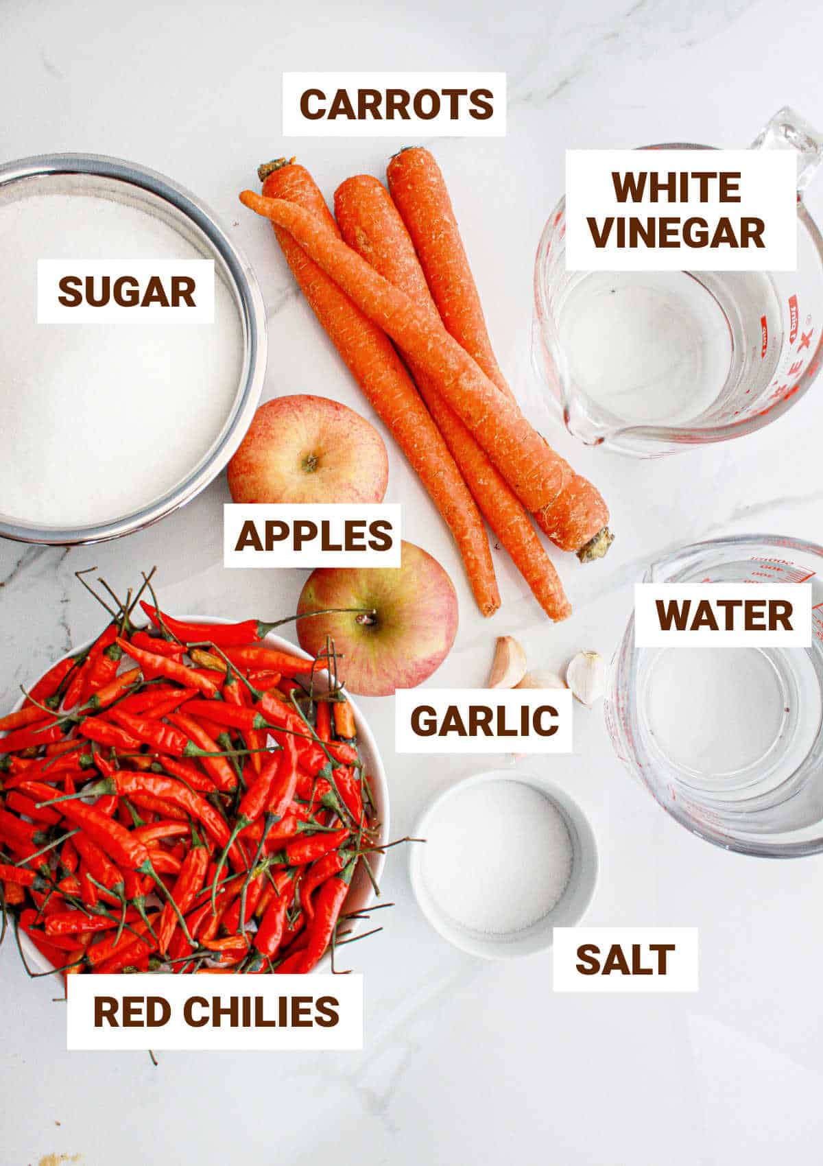 White surface with bowls containing ingredients for red chili jam including apples, carrots and garlic.