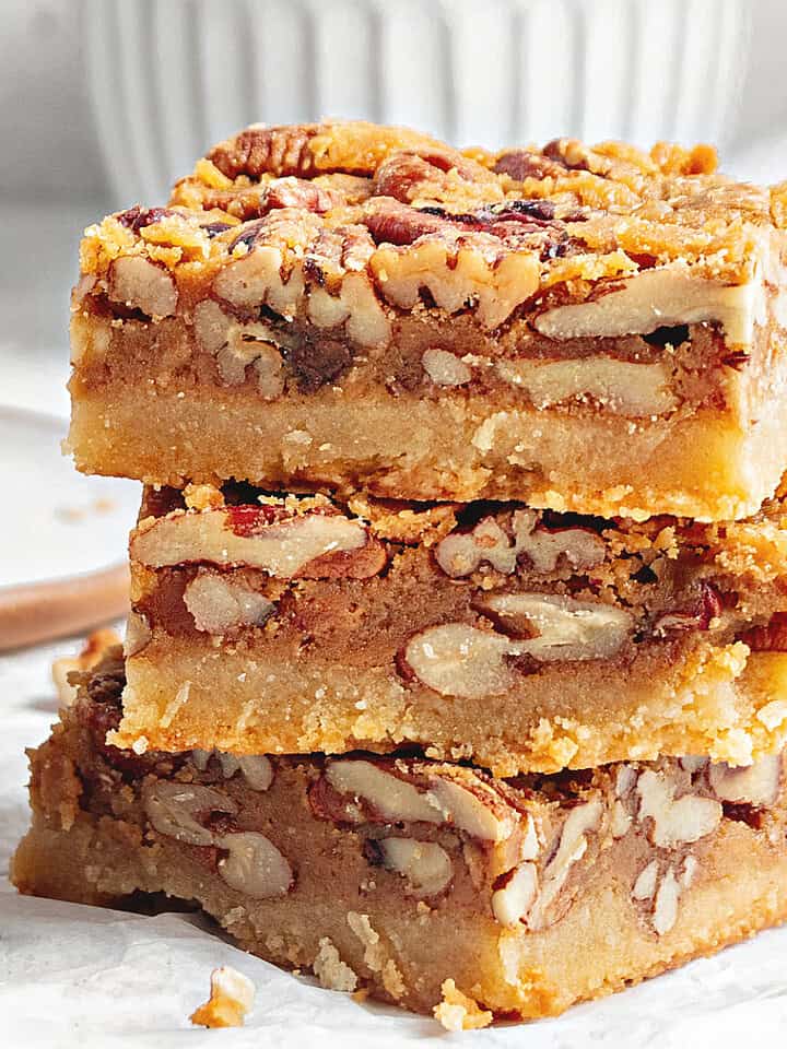 Three pecan bars stacked on white paper. White gray bowl in the background.