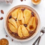 Top view of pear upside down cake on a white plate. Pecans, coffee cups, silver forks. Whitish background.