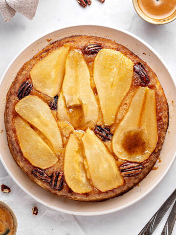 Top view of pear upside down cake on a white plate. Pecans, coffee cups, silver forks. Whitish background.