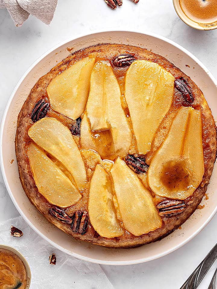 Top view of pear upside down cake on a white plate. Pecans, coffee cups, silver forks. Whitish background.