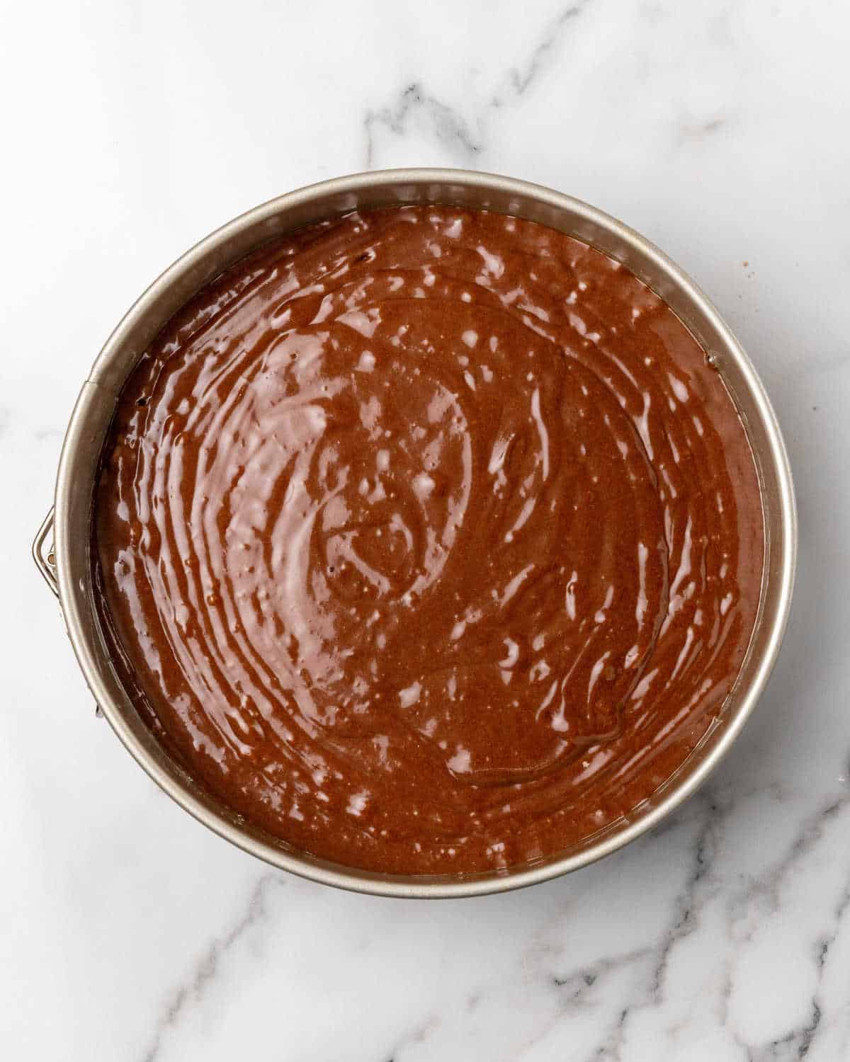 Metal round cake pan with chocolate cake batter on a white marble surface. 