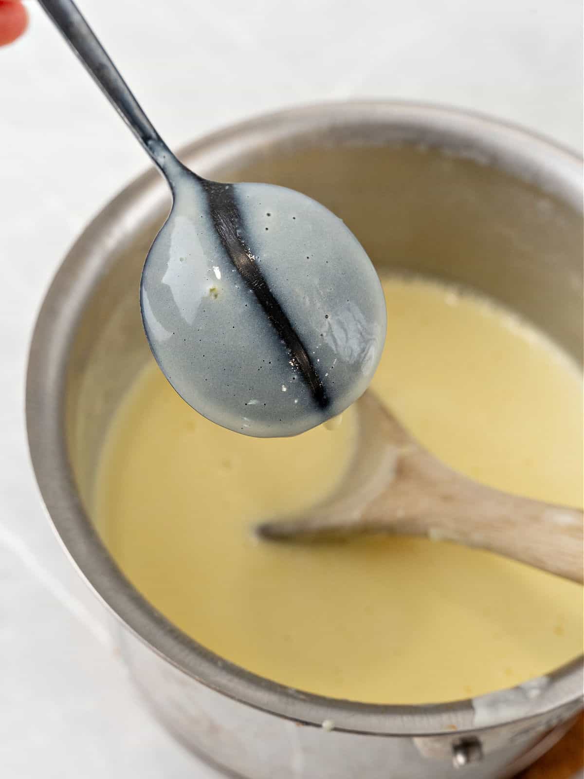 Dark spoon with custard over saucepan with rest of mixture. Light gray background.