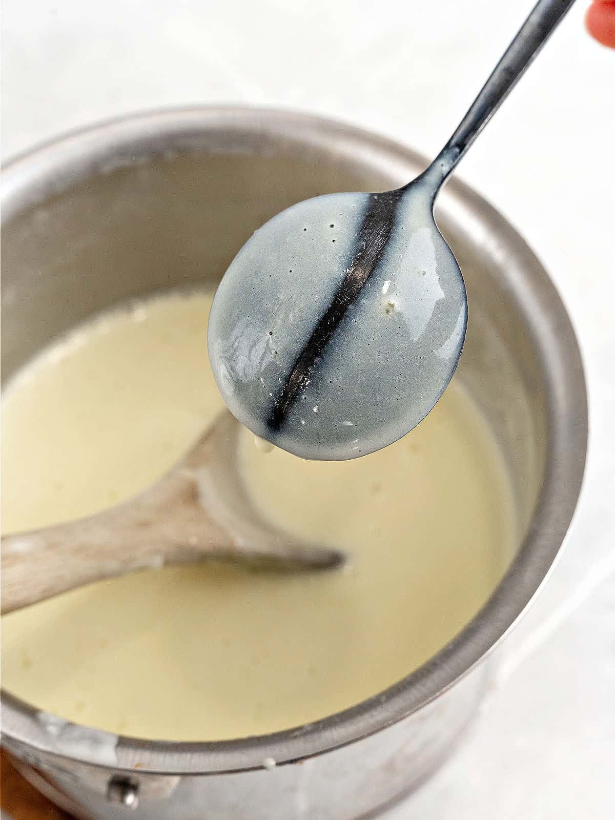 Dark spoon with custard over saucepan with rest of mixture. White background.
