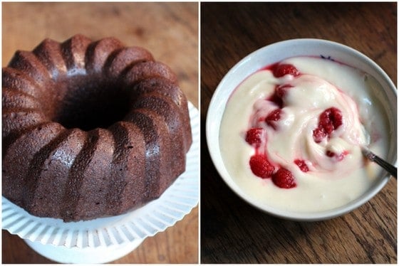 A chocolate bundt cake on a white cake stand and a bowl of glaze with raspberries