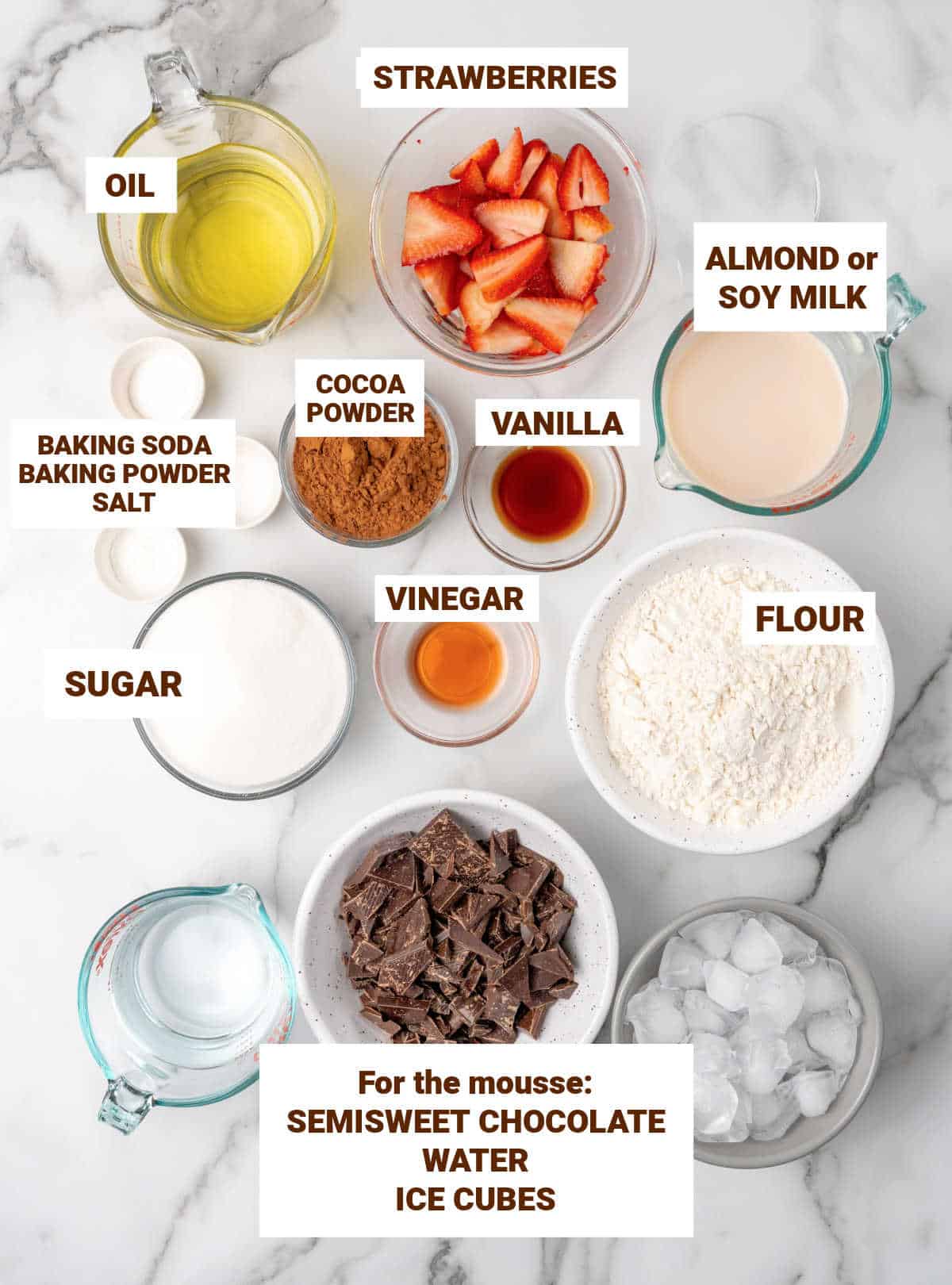 Bowls on white marble containing ingredients for vegan chocolate cake including strawberries, oil, flour, cocoa powder, vegetable milk, sugar, water.