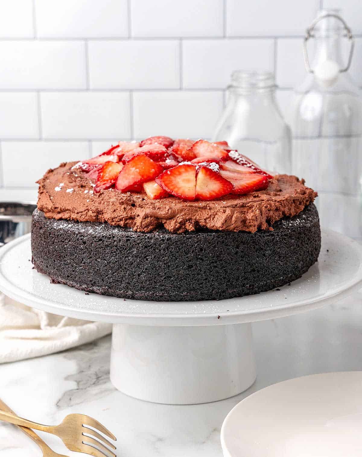 White cake stand with chocolate mousse cake topped with strawberries. White tile background.