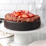 A strawberry topped chocolate mousse cake on a white cake stand. White tile background.
