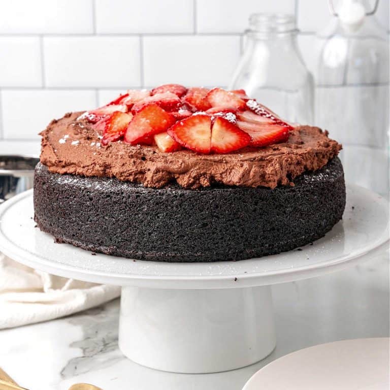 A strawberry topped chocolate mousse cake on a white cake stand. White tile background.