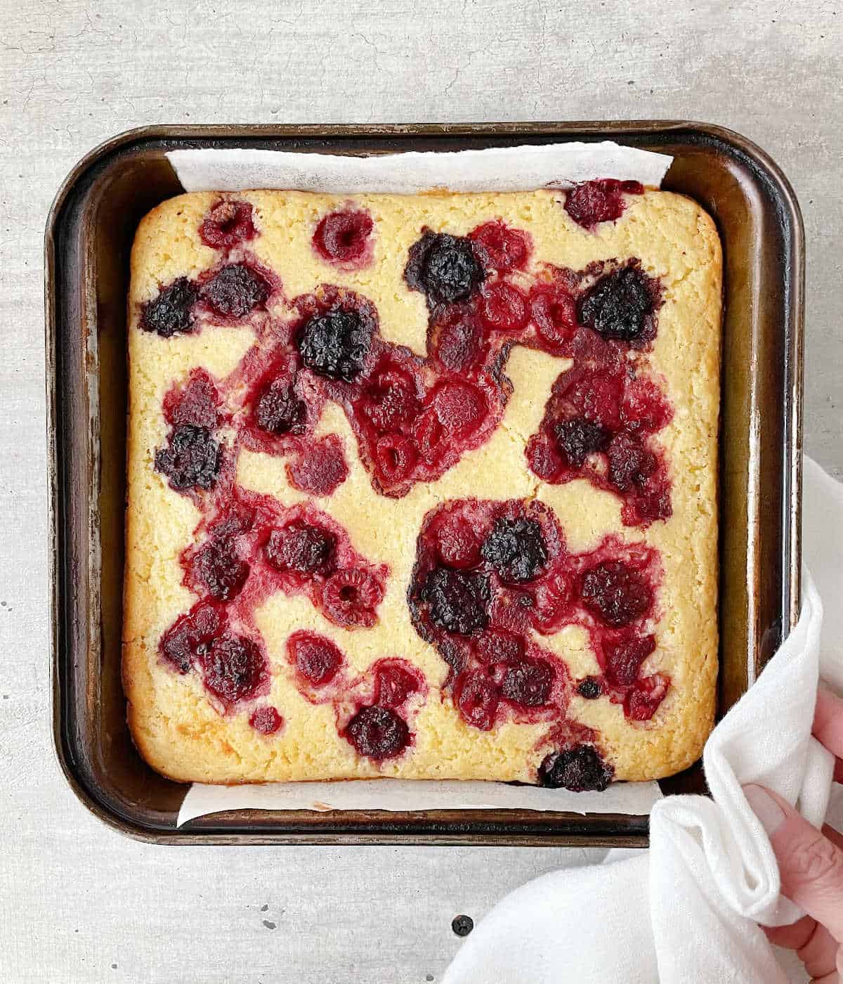 Hand holding baked ricotta cake with berries on dark metal square pan on grey surface.