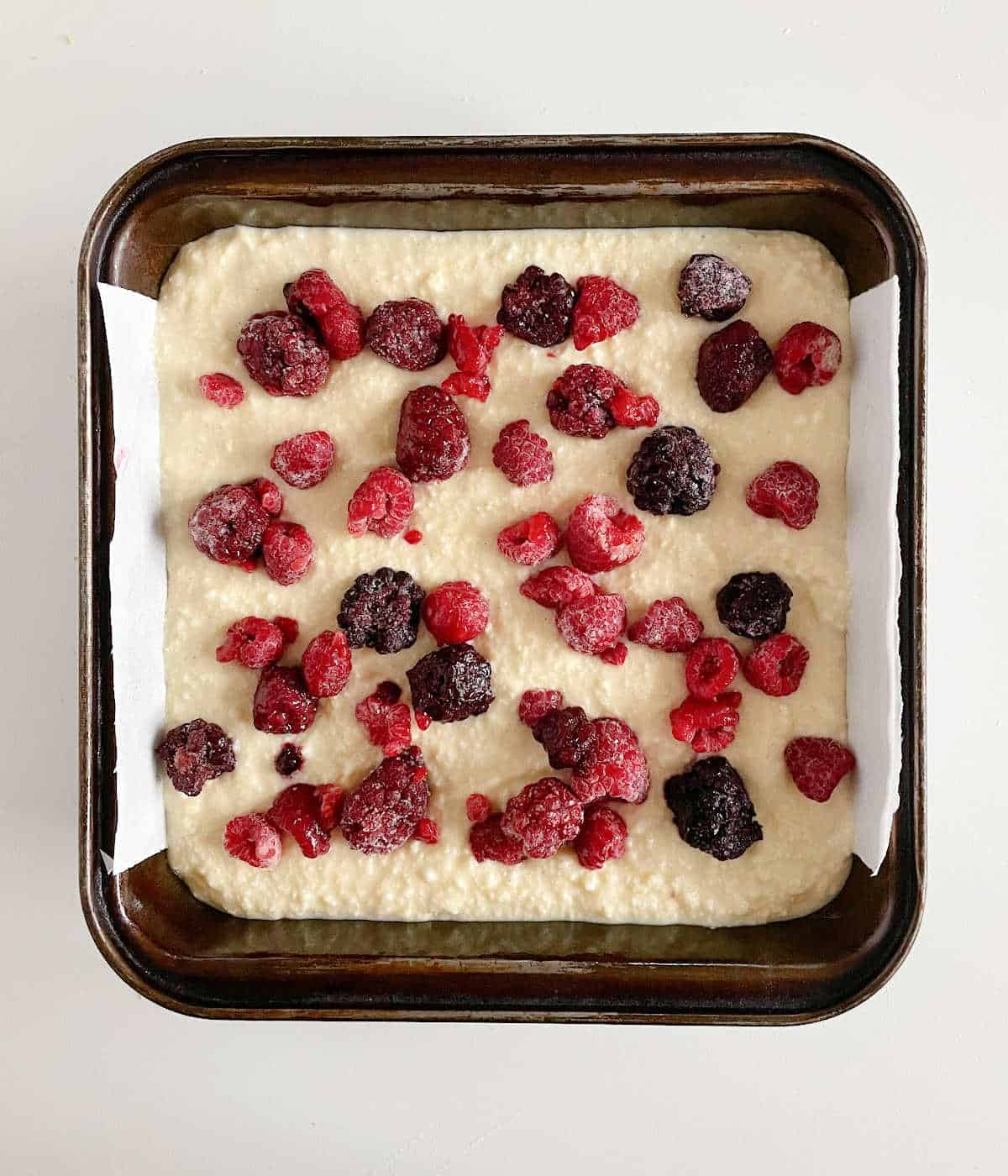 Dark metal square pan on grey surface with unbaked berry ricotta cake mixture.