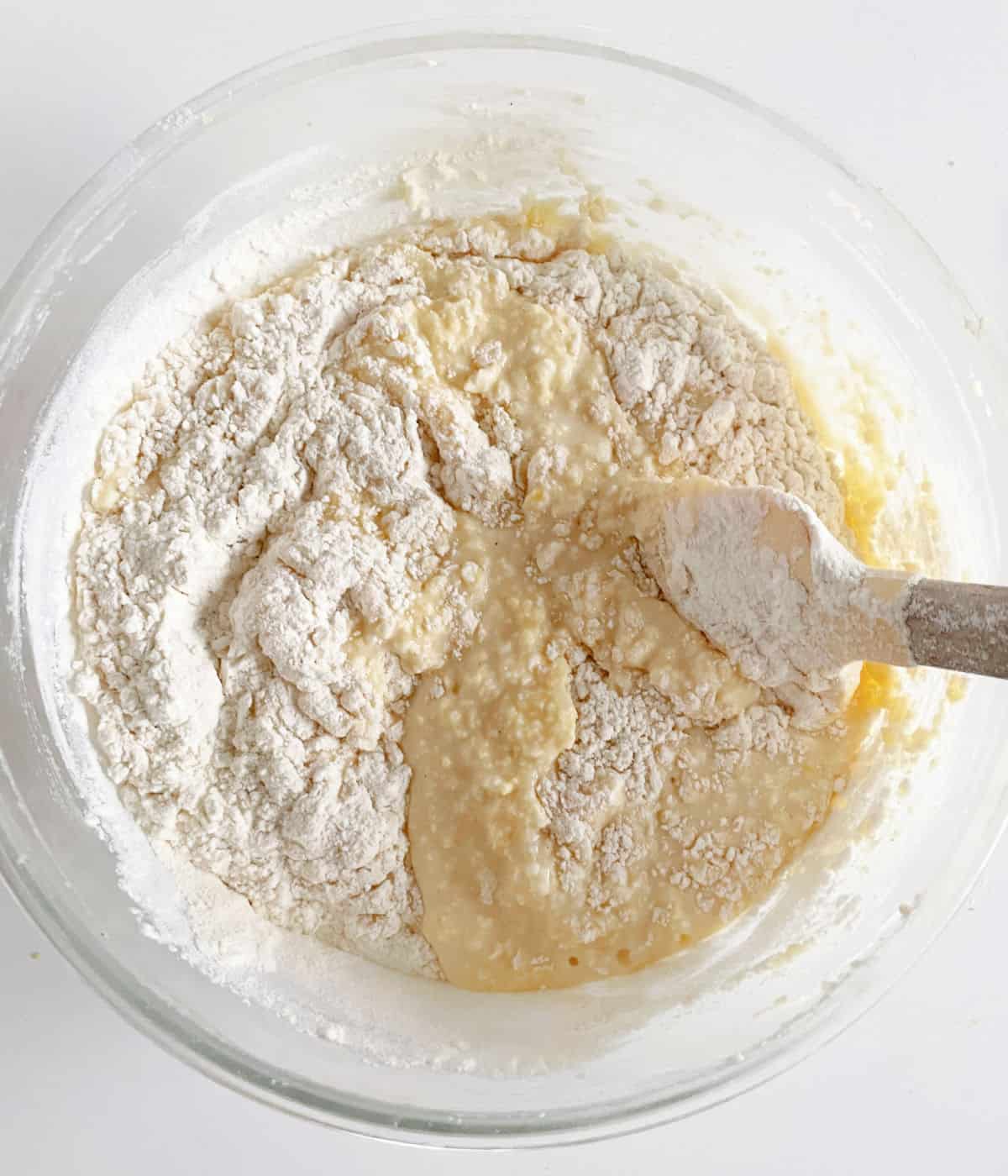 Flour being mixed into ricotta cake mixture with spatula. Glass bowl on a white table. 