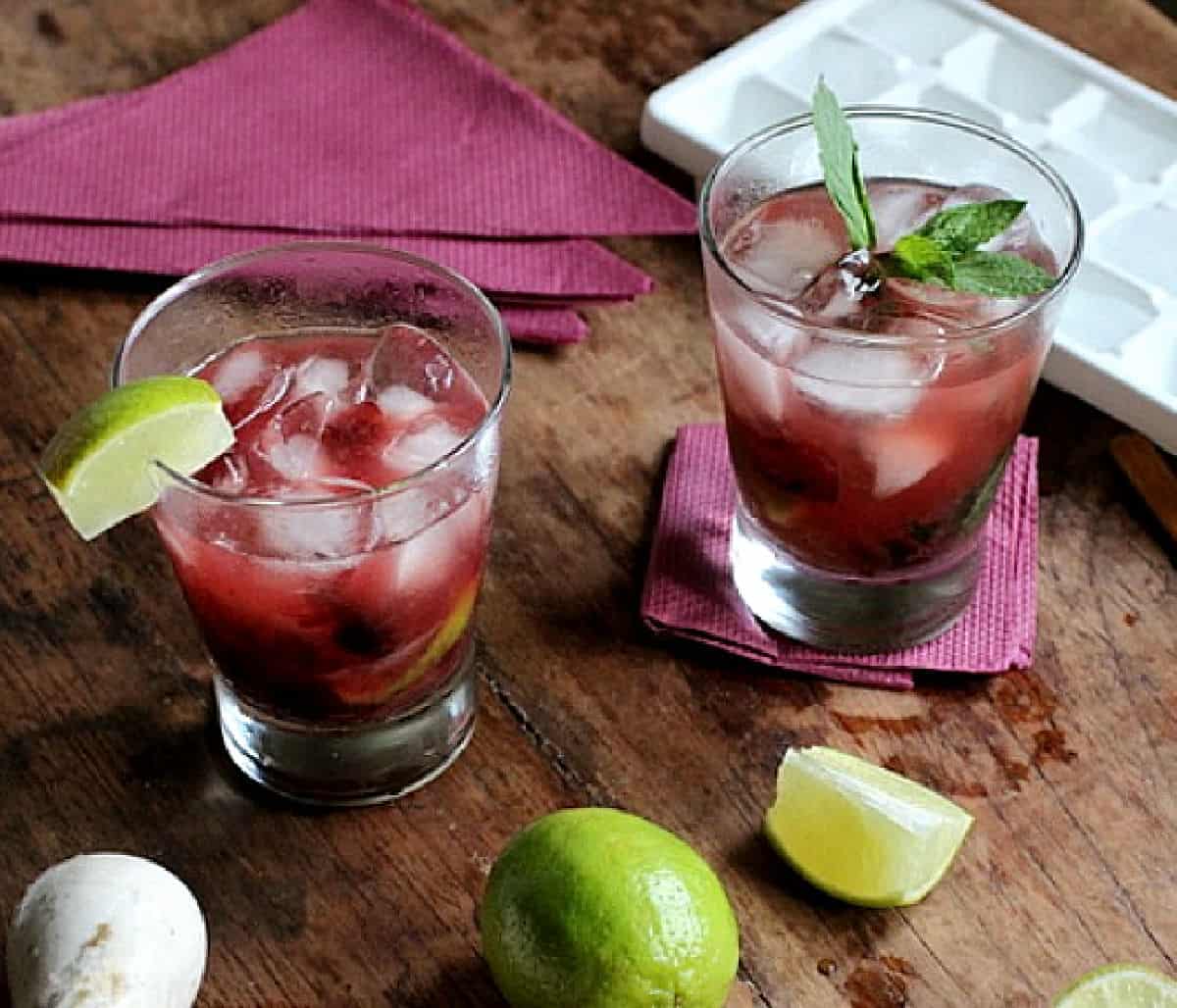 Two glasses with cherry cocktail on wooden table, limes, purple napkins.