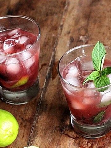 Wooden table with glasses of cherry cocktail, limes and mint sprigs.