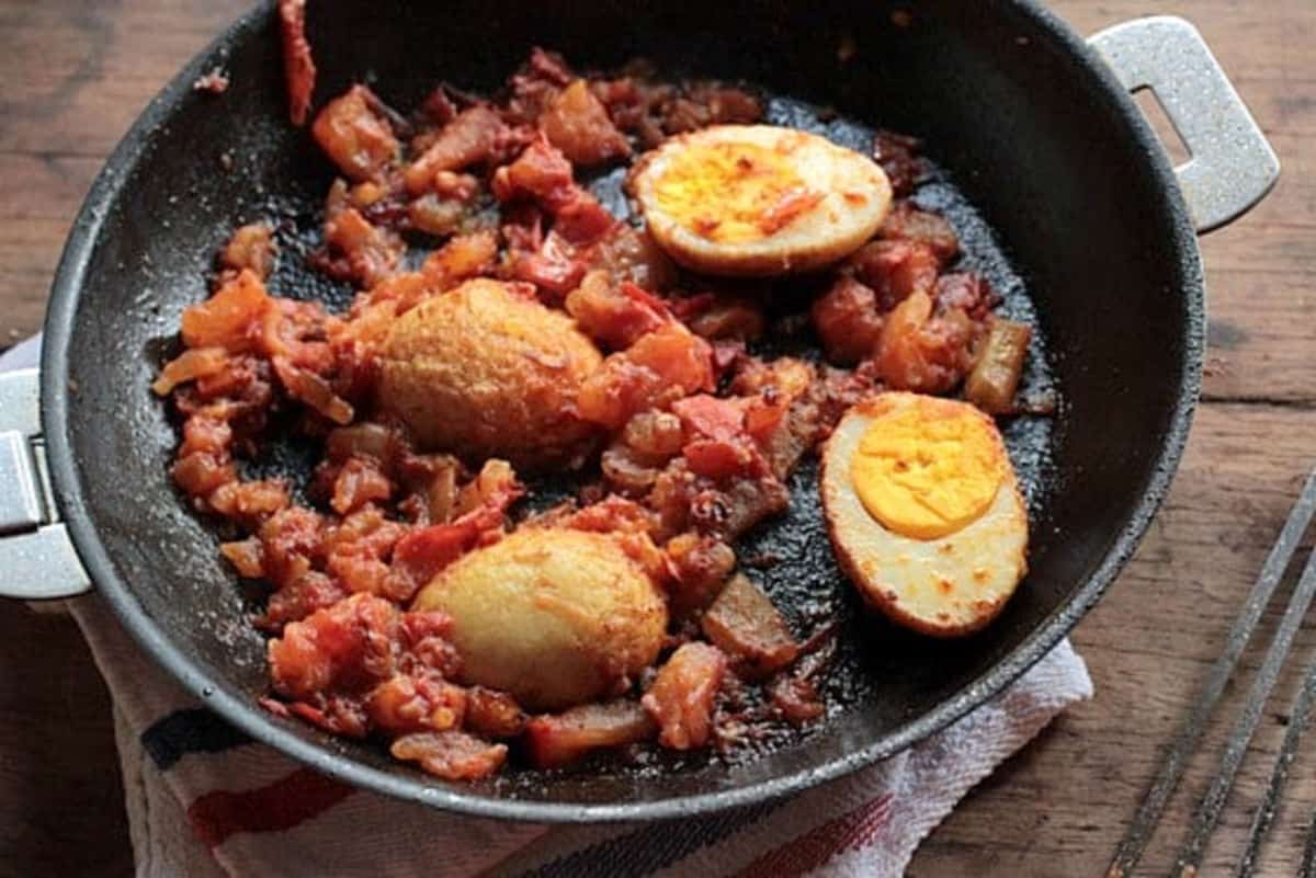 Top view of fried hard boiled eggs and tomato sauce in a dark metal skillet on a wooden table. 