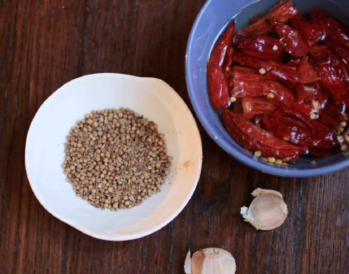 Wooden table with bowl containing ingredients for harissa including seeds, garlic cloves, and dried peppers.