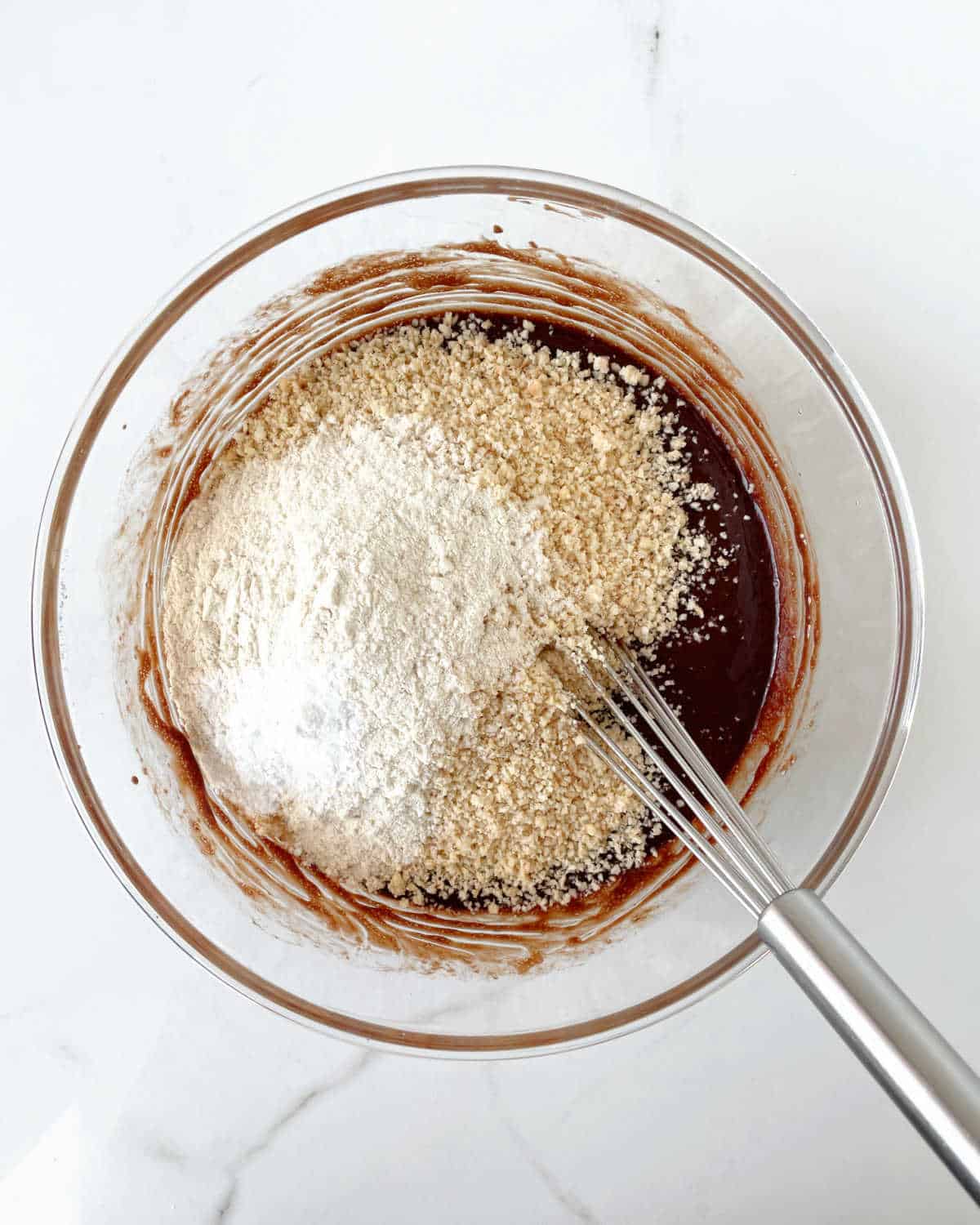 Ground hazelnuts and flour on a glass bowl with brownie batter on a white marble surface.