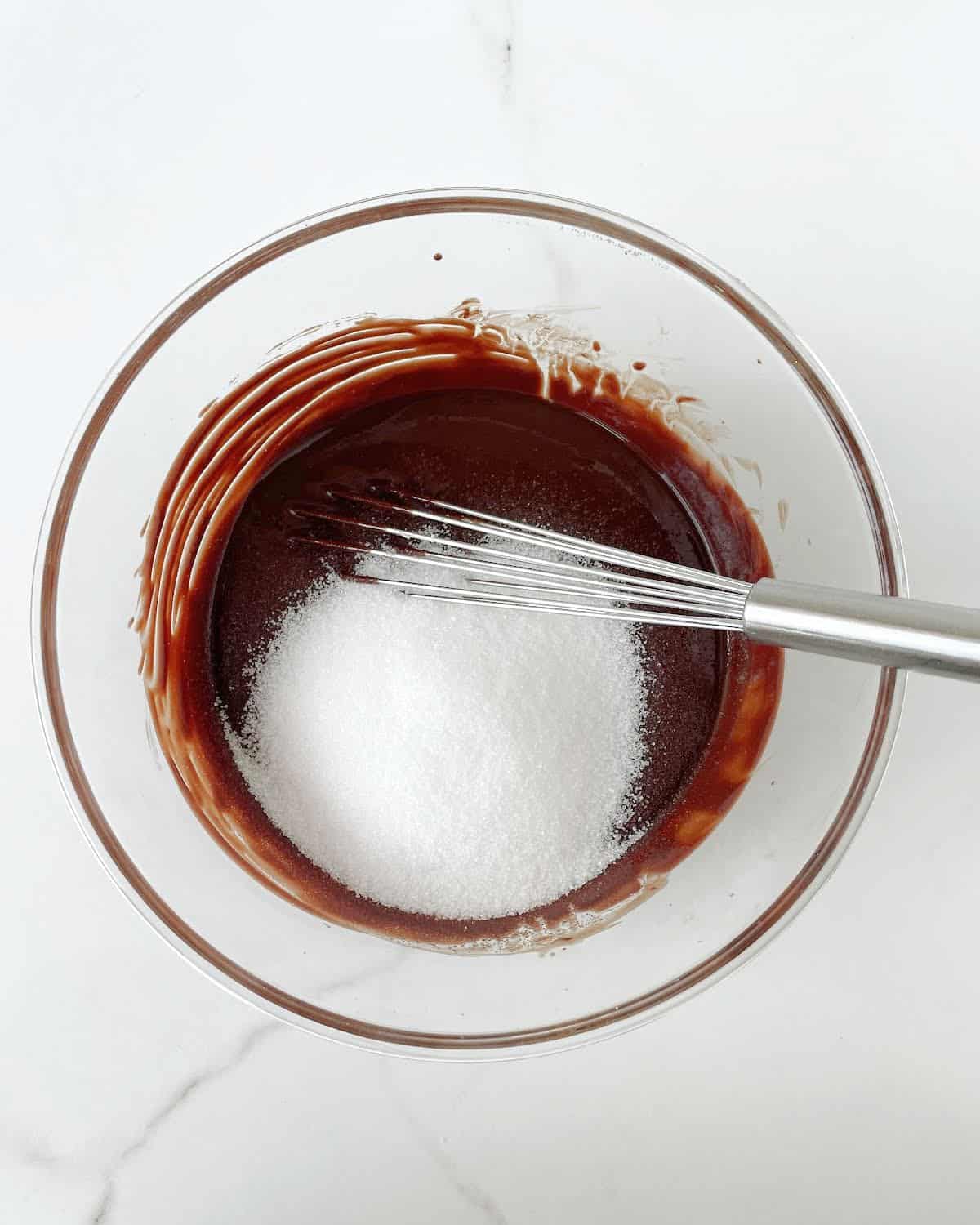 Sugar added to brownie batter in a glass bowl with a whisk inside. White marble surface. 