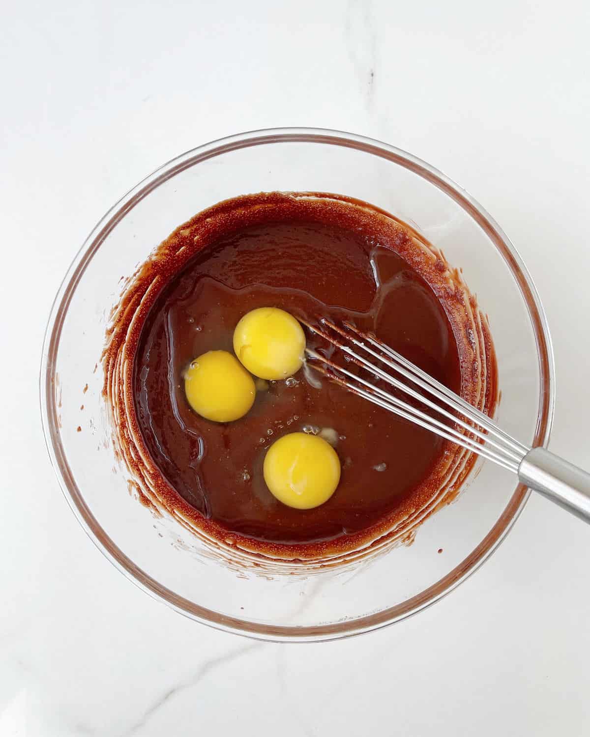 Three eggs added to brownie batter in a glass bowl. A whisk inside. White marble surface.