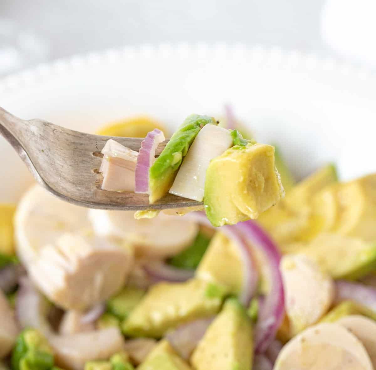 Fork with bite of avocado, heart of palm and red onion salad. White bowl with salad beneath. 