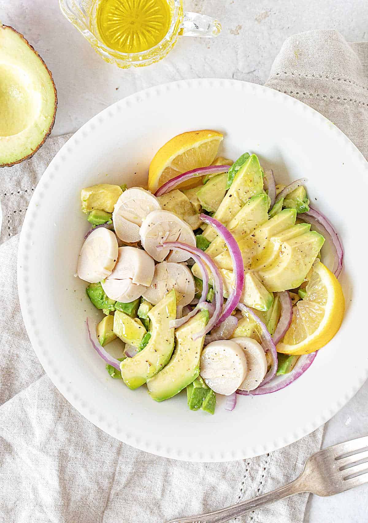 Grey cloth and surface with white plate containing avocado, red onion, and heart of palm salad. Olive oil jar.