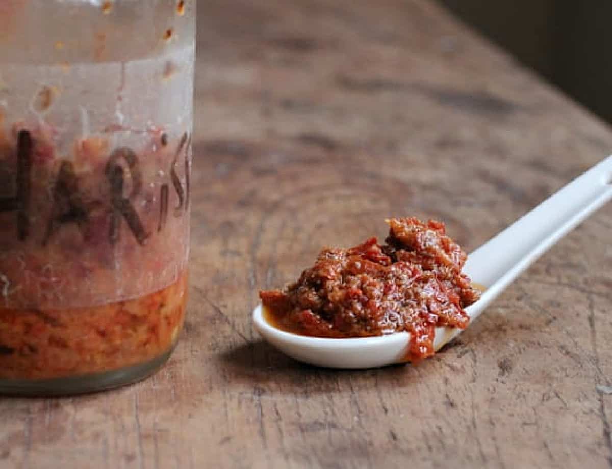 Glass jar and white spoon with harissa paste on a wooden table. 