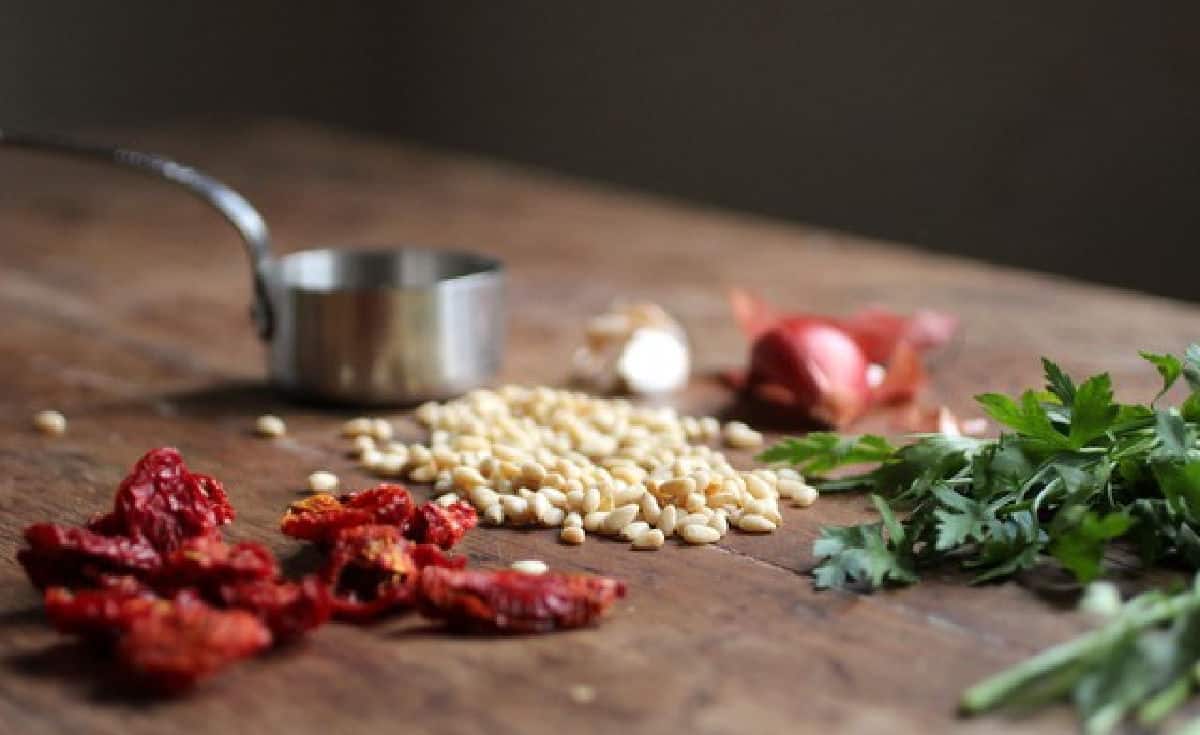 Wooden table with ingredients for sun dried tomato pesto including shallots, pine nuts, and parsley.