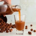 Close up pouring hazelnut liqueur into shot glass. Beige gray background, whole hazelnuts.