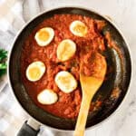 Golden eggs with tomato sauce in a dark skillet. Wooden spoon. White checkered background.