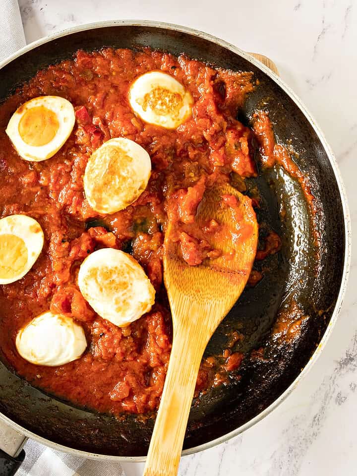 Golden eggs with tomato sauce in a dark skillet. Wooden spoon. White checkered background.