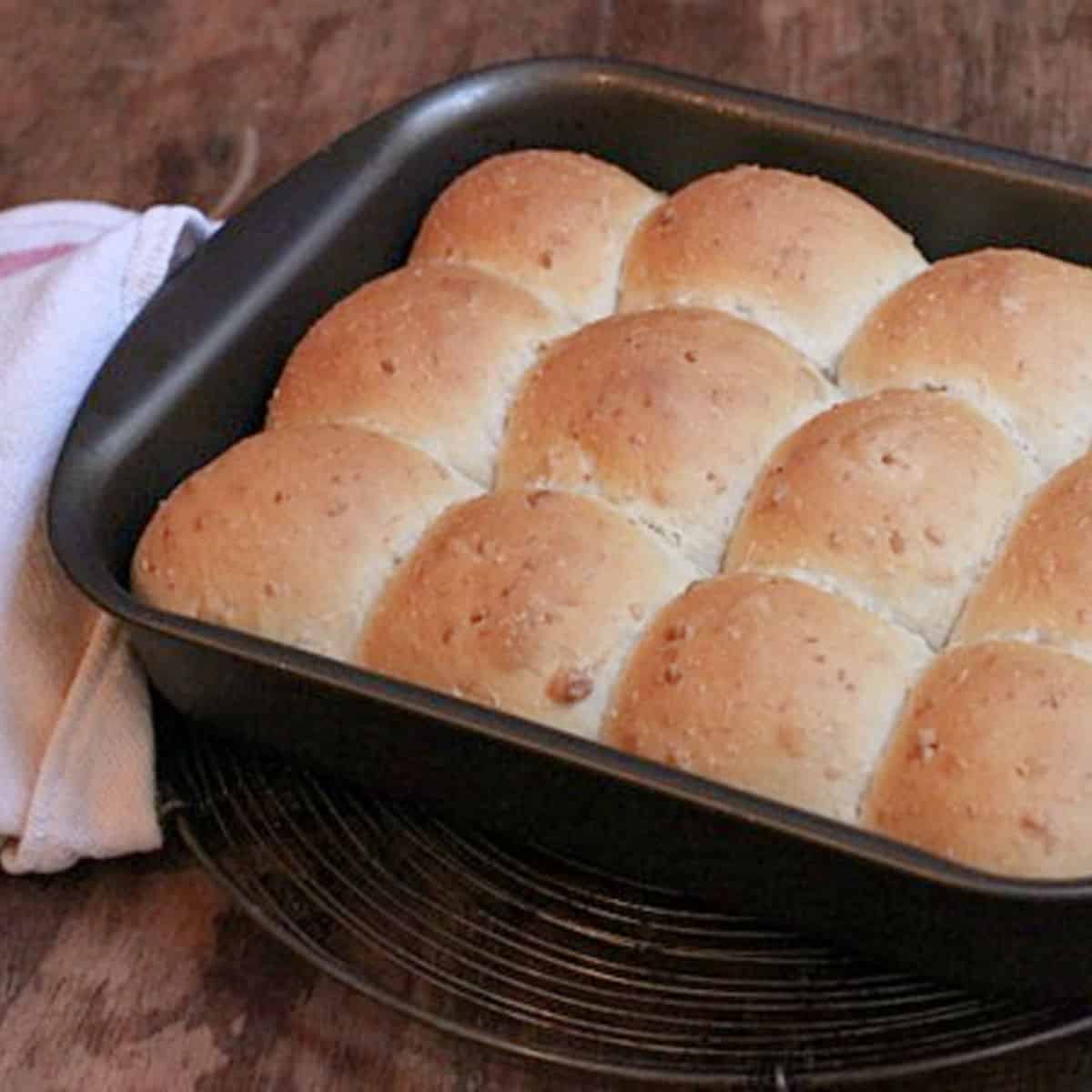 Close up of baked coconut buns in a dark metal pan with kitchen towel on a wooden table.