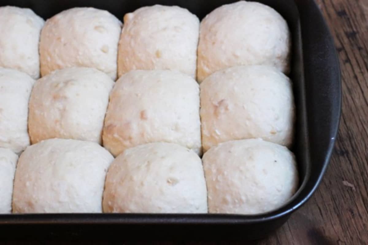 Raw and risen coconut buns in a dark metal pan on a wooden table. 