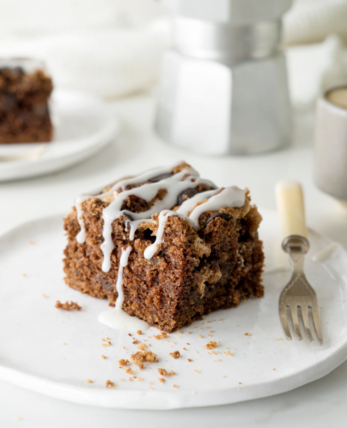 Square of glazed chocolate coffee cake on a white plate with fork. White background with coffee pot.
