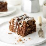 Single glazed chocolate cake square on a white plate, a fork. White background with metal coffee pot and more cake.
