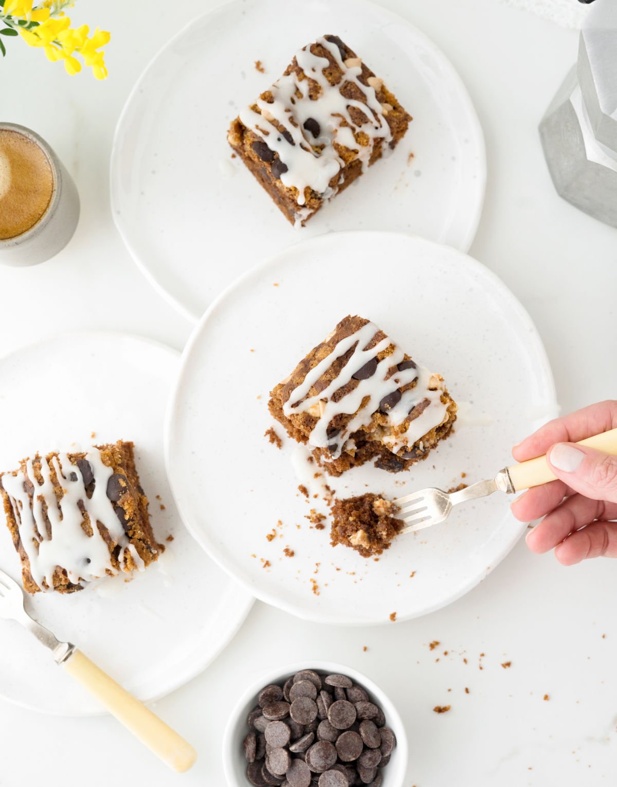 Overview of three white plates with squares of glazed chocolate cake. White surface. A hand with a bite on a fork. 